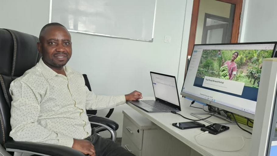 Smiling Black man in an office, seated at a desk with a laptop and a monitor displaying the 'Tearfund Brand Portal' with a person holding bananas.