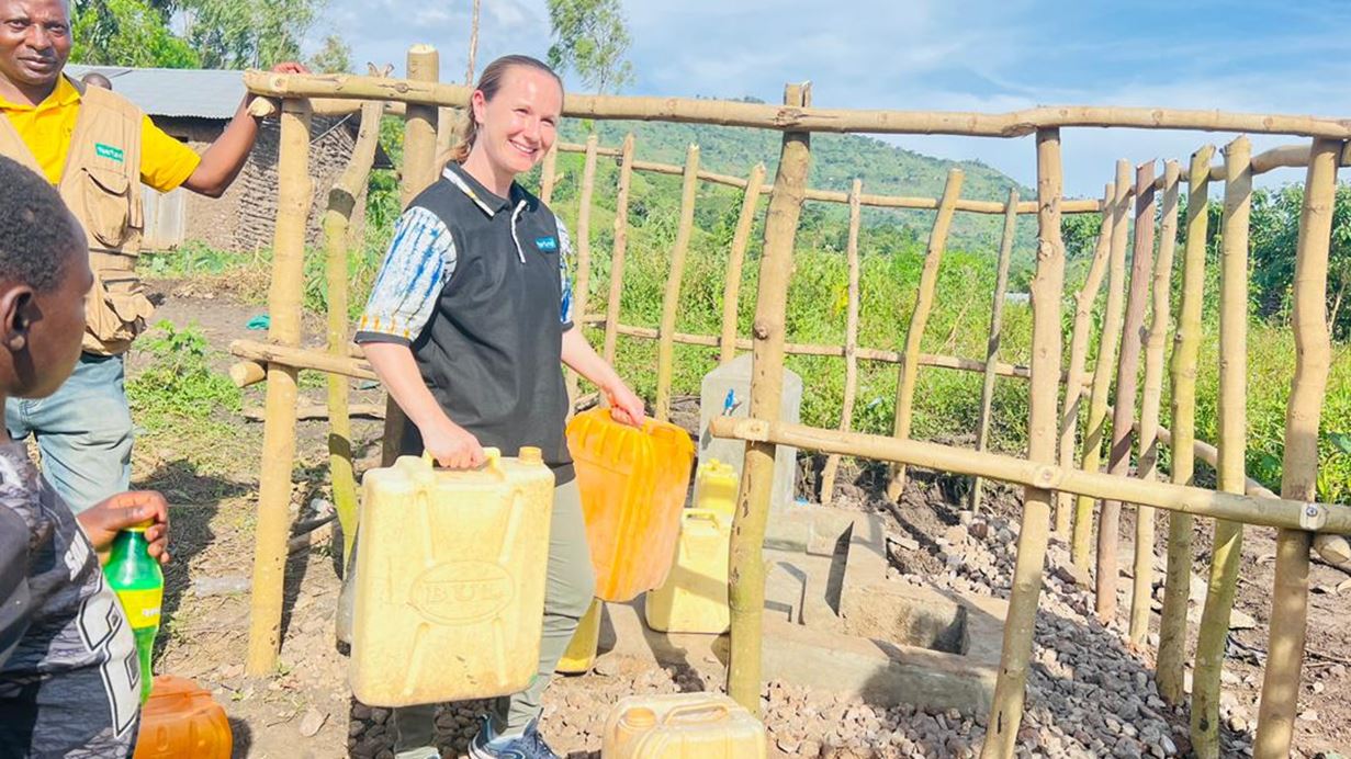 A woman smiles, holding two yellow jerry cans at an outdoor water point with a wooden fence. A man in a vest and a child are nearby, with green hills in the background.