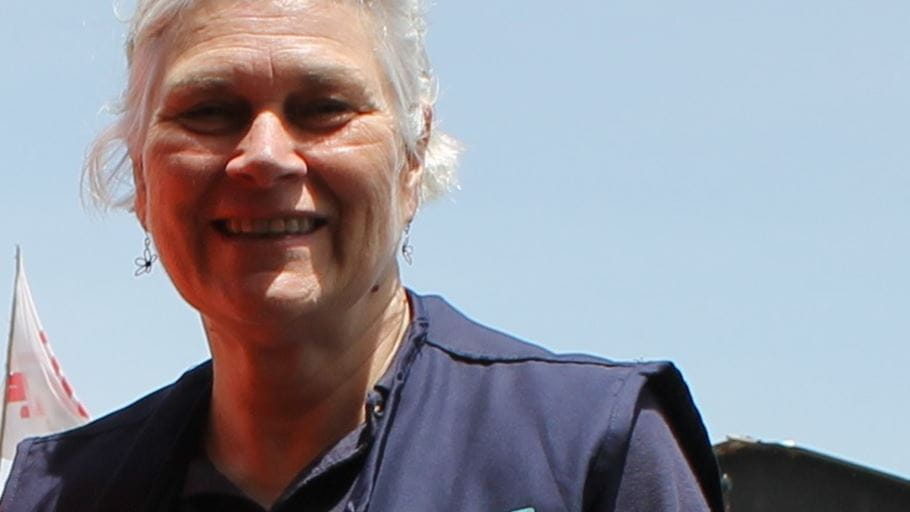 Close-up of a smiling woman with grey hair and a dark blue top against a bright sky, with a white and red flag visible on the left.