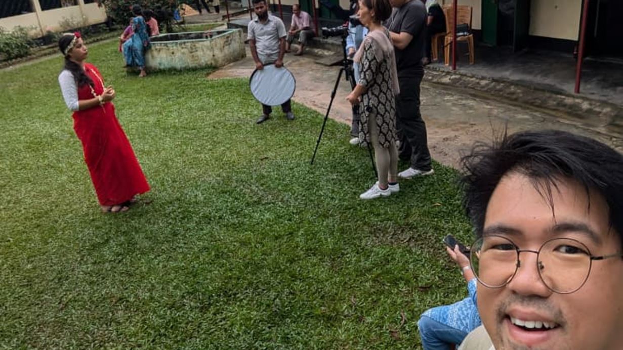 A man in glasses takes a selfie, capturing a photoshoot in progress on a grassy area. A woman in a red traditional dress and flowers in her hair poses, while a man holds a light reflector and another person adjusts a camera on a tripod. Other people are visible in the background.