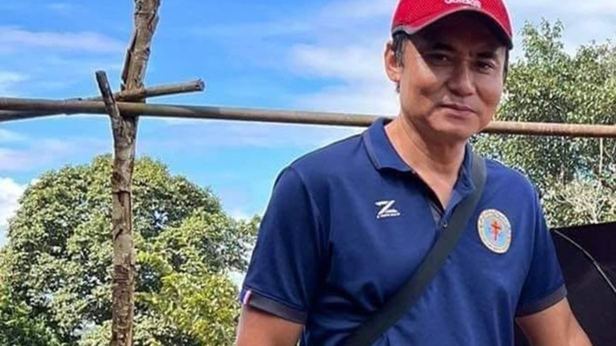 Man in a red cap and blue polo shirt with a red cross badge, looking forward. Behind him are green trees and a wooden cross-shaped structure against a blue sky.