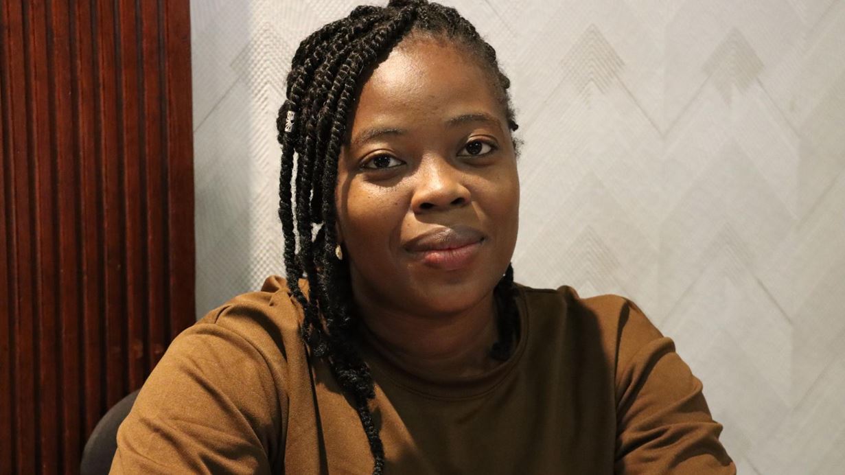 Close-up of a woman with dark skin and braided hair, wearing a brown top, looking directly ahead with a slight smile.