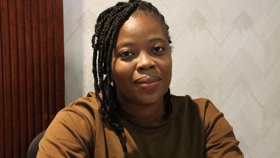 Close-up of a woman with dark skin and braided hair, wearing a brown top, looking directly ahead with a slight smile.