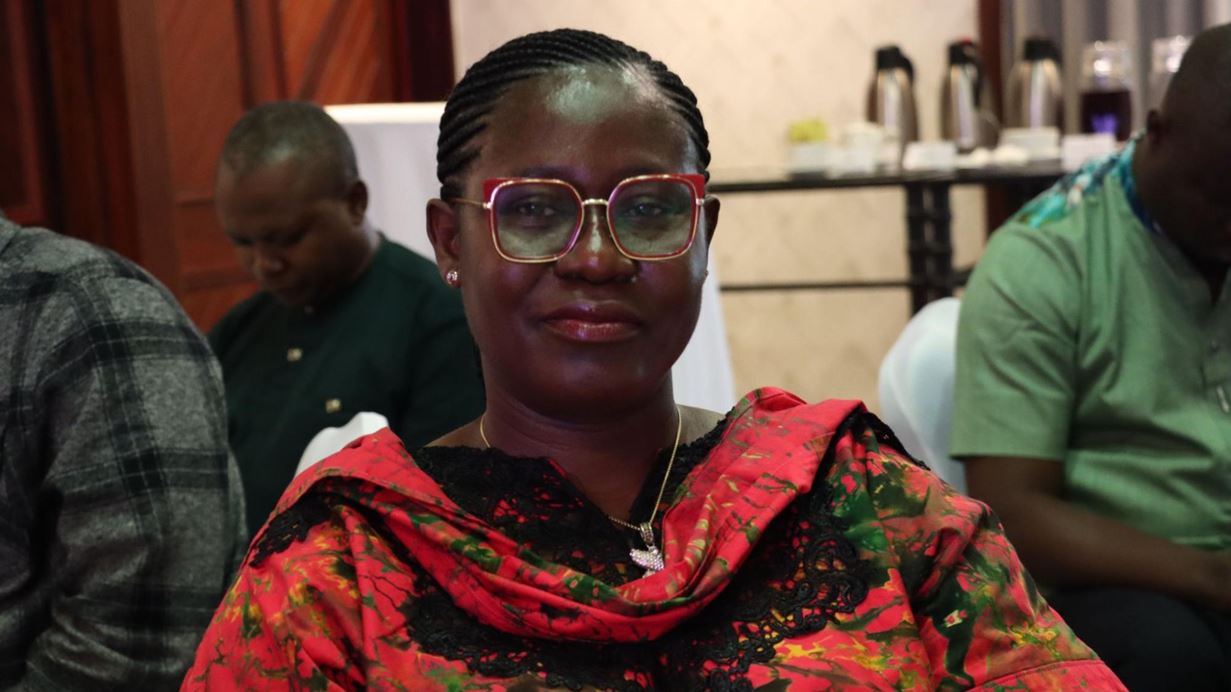 A woman with braided hair and red-framed glasses smiles while wearing a red and black patterned top. Other people are blurred in the background.
