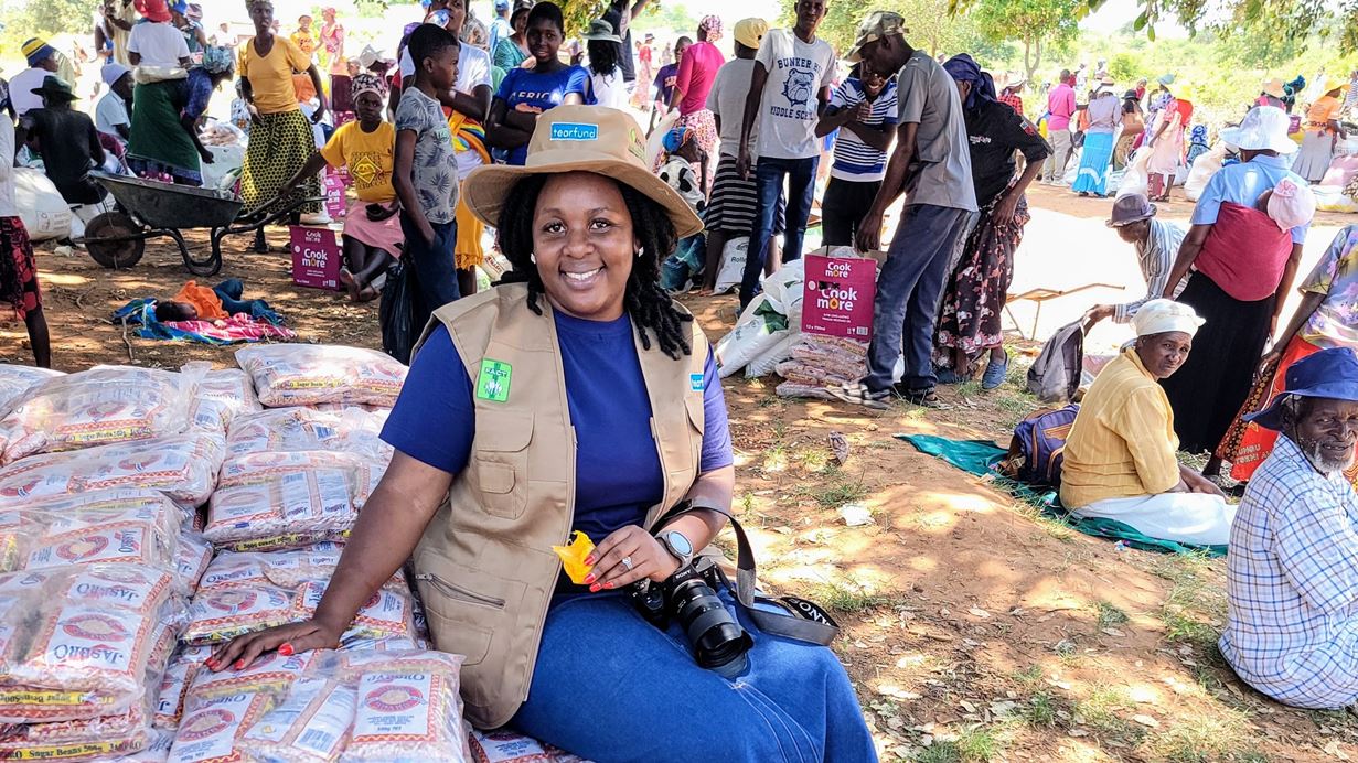 Smiling woman in a hat and vest sitting next to stacks of bagged goods, with a large crowd of people in the busy background.