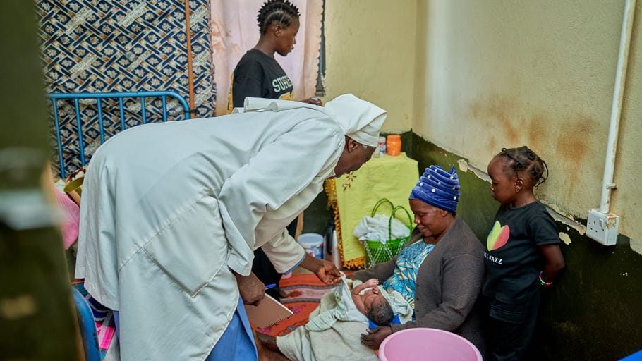A healthcare worker in a white uniform leans over a newborn baby, held by a seated woman in a blue headscarf. A young girl and another woman observe the scene.