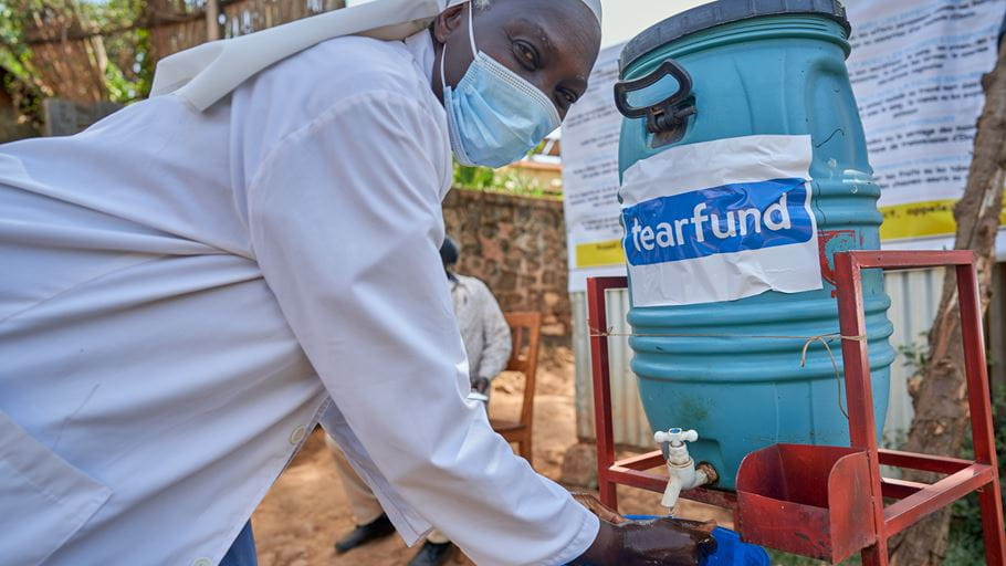 A man wearing a white shirt and surgical mask washes his hands at an outdoor handwashing station. A blue barrel labelled 'tearfund' provides water from a tap.