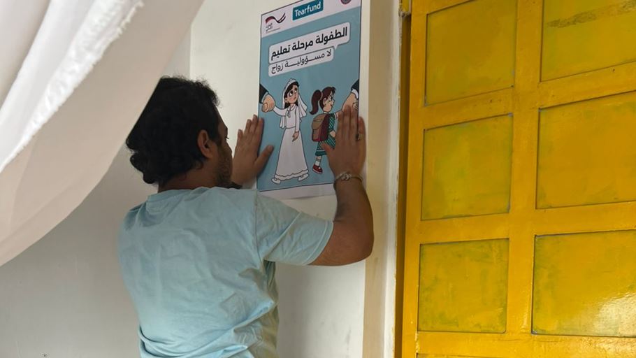 A man in a light blue t-shirt presses a white poster onto a wall. The poster, featuring the Tearfund logo, shows two girls being pulled by hands: one in a white wedding dress and another in a school uniform. Visible Arabic text translates to 'Childhood is a stage of education, not a responsibility of marriage'. A yellow door is to the right, and a white curtain hangs above.