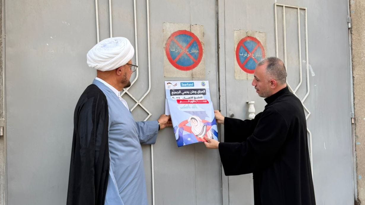 Two men are attaching a poster to a grey metal door. The man on the left wears a white turban and robes, while the man on the right wears dark clothing. The poster, which includes Arabic text and a 'Tearfund' logo, depicts a graphic of a person surrounded by hands. Faded red circular signs, resembling 'no waiting' or 'no parking' indicators, are on the door above.