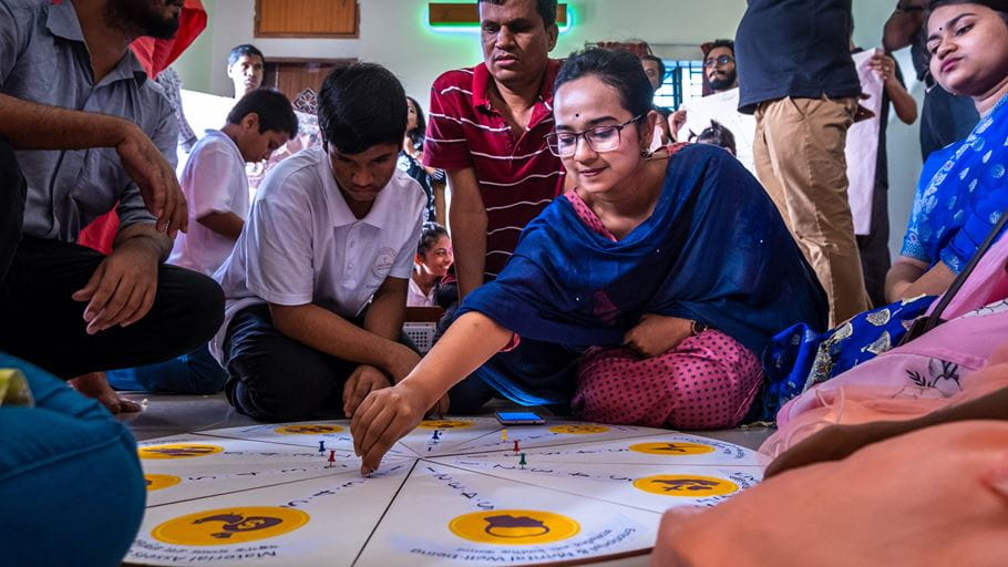 A young woman in glasses and a blue shawl places a pin on a large circular board game on the floor, surrounded by other adults and young people.