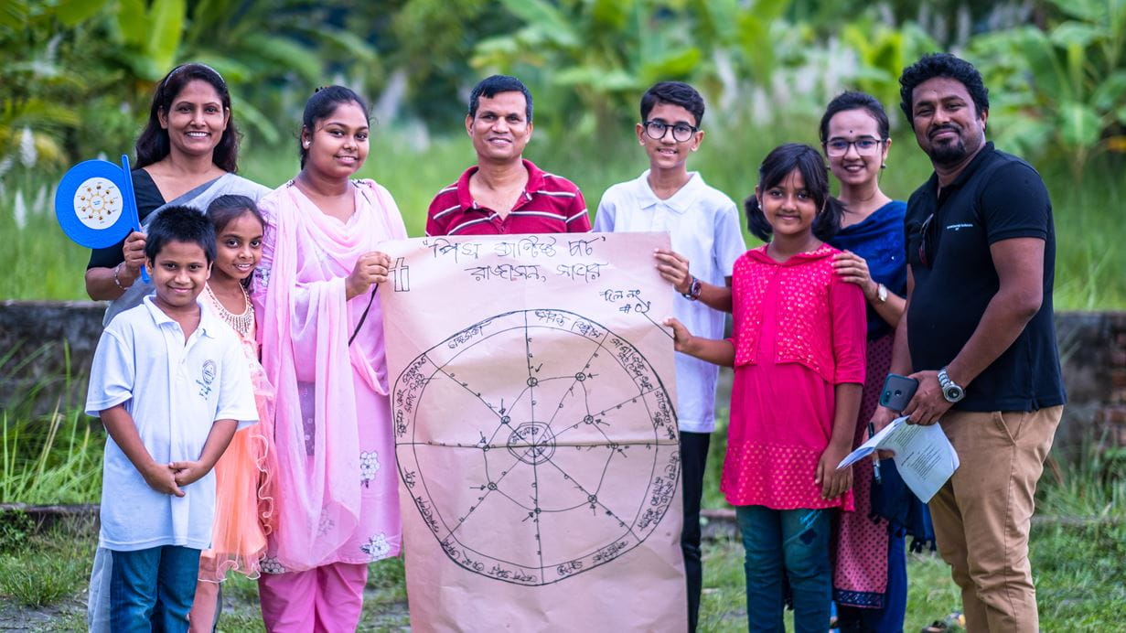 A smiling group of nine people, including adults and children, stand outdoors against a green background. Two adults in the centre hold up a large piece of brown paper with a hand-drawn circular diagram and text. A woman on the left holds a blue, circular device with lights, and a man on the right holds white papers.