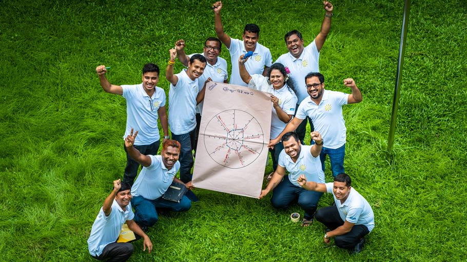 Ten adults, mostly men, wearing light polo shirts with yellow logos, stand and crouch on vibrant green grass. They are smiling, around a large brown sheet of paper displaying a circular diagram.