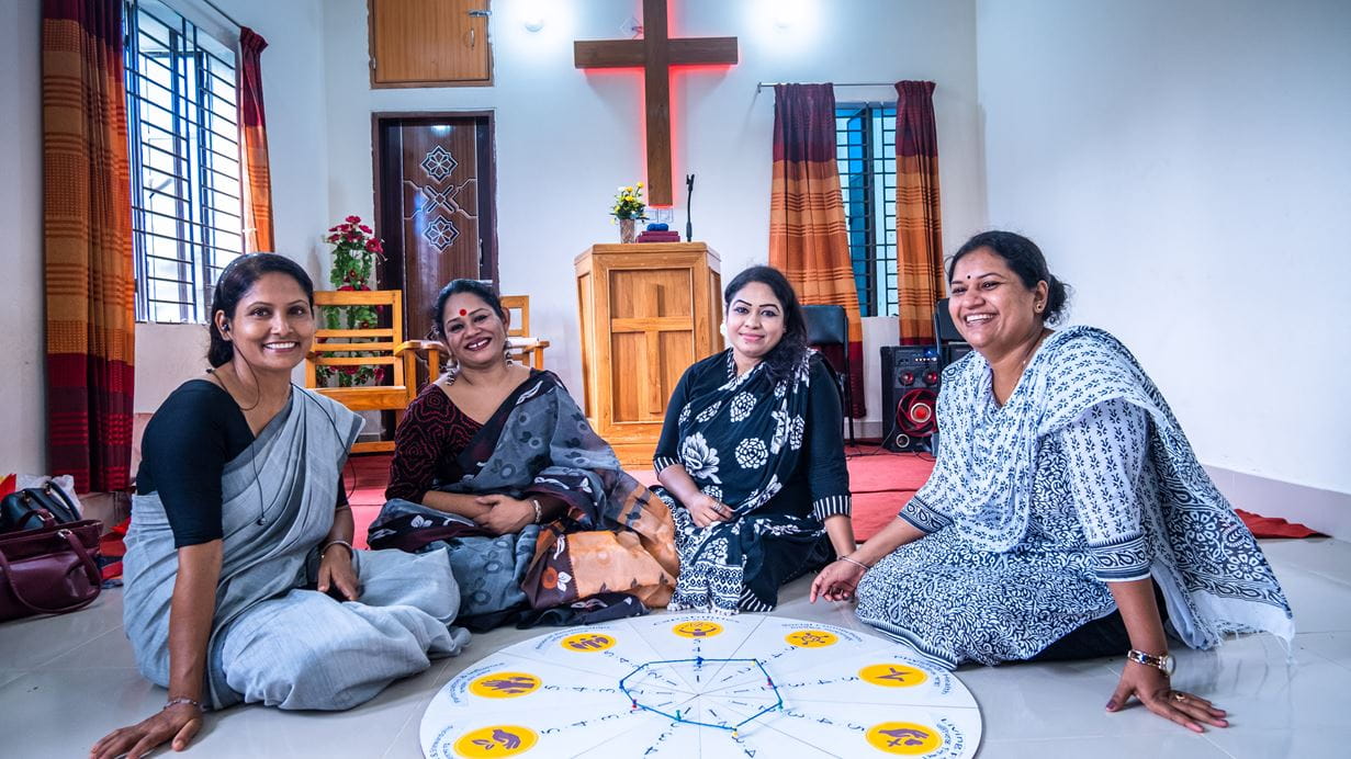 Four smiling women sit on the floor in a room, with a large circular board in front of them and a wooden cross and pulpit in the background.