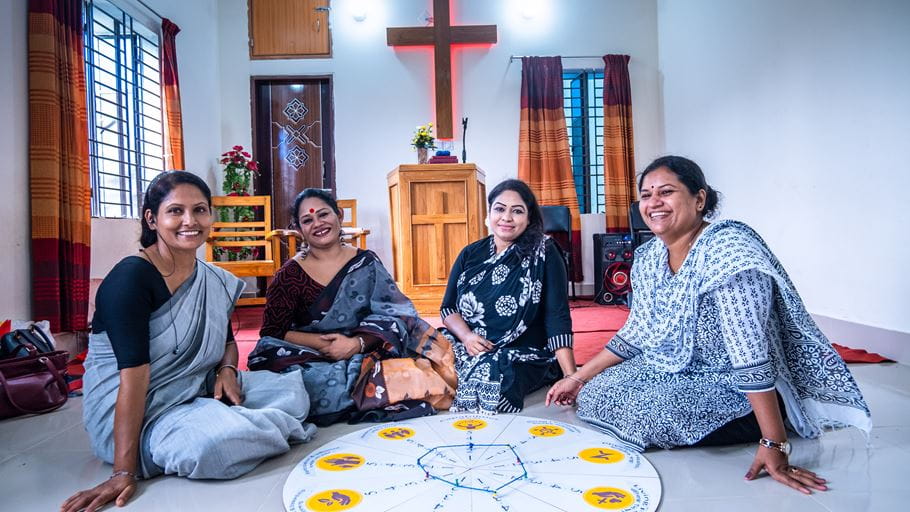 Four smiling women sit on the floor in a room, with a large circular board in front of them and a wooden cross and pulpit in the background.