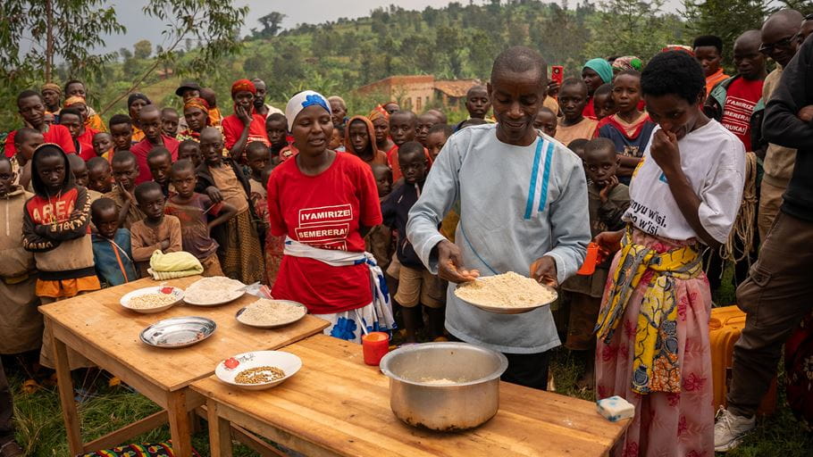 A man presents a plate of pale flour at an outdoor wooden table, surrounded by a crowd of people, including many children, on a green hillside. The table displays various grains, flours, and a cooking pot. A woman in a red t-shirt stands behind the table.