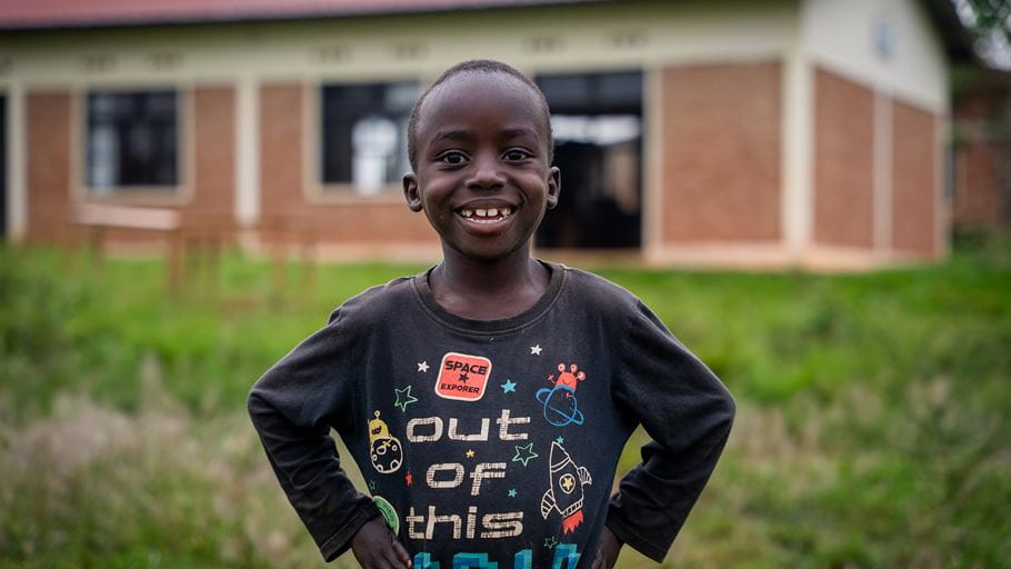 Young Black boy smiling broadly, wearing a dark long-sleeved t-shirt with colourful space-themed designs. He stands outdoors with hands on hips, a blurred building and green grass in the background.