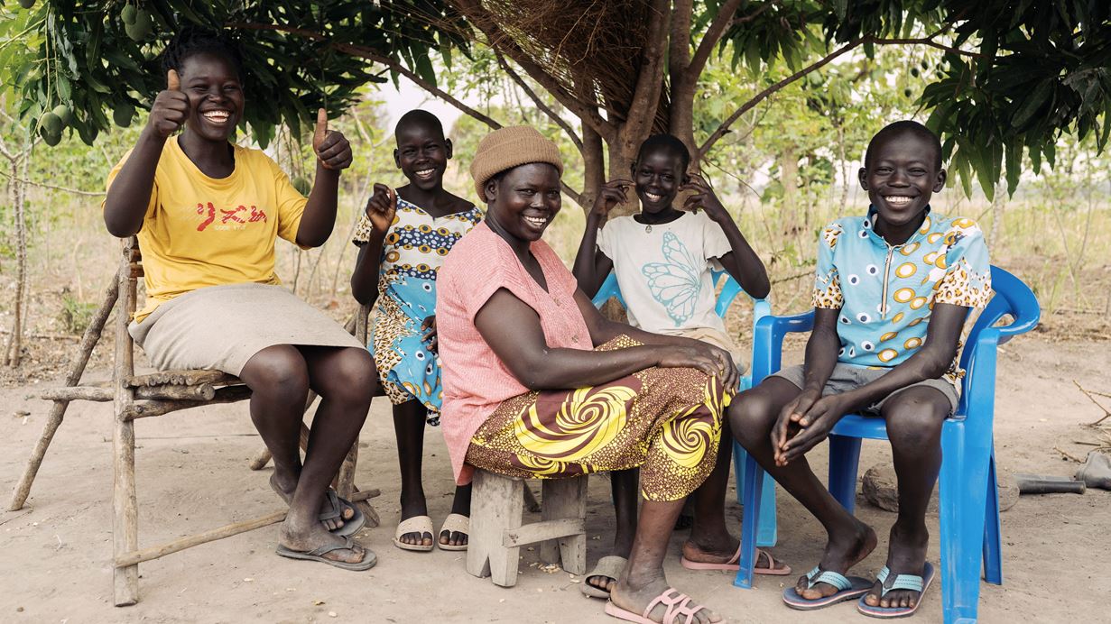 Mary and her children smile as they sit together on chairs outside under the shade of a big tree