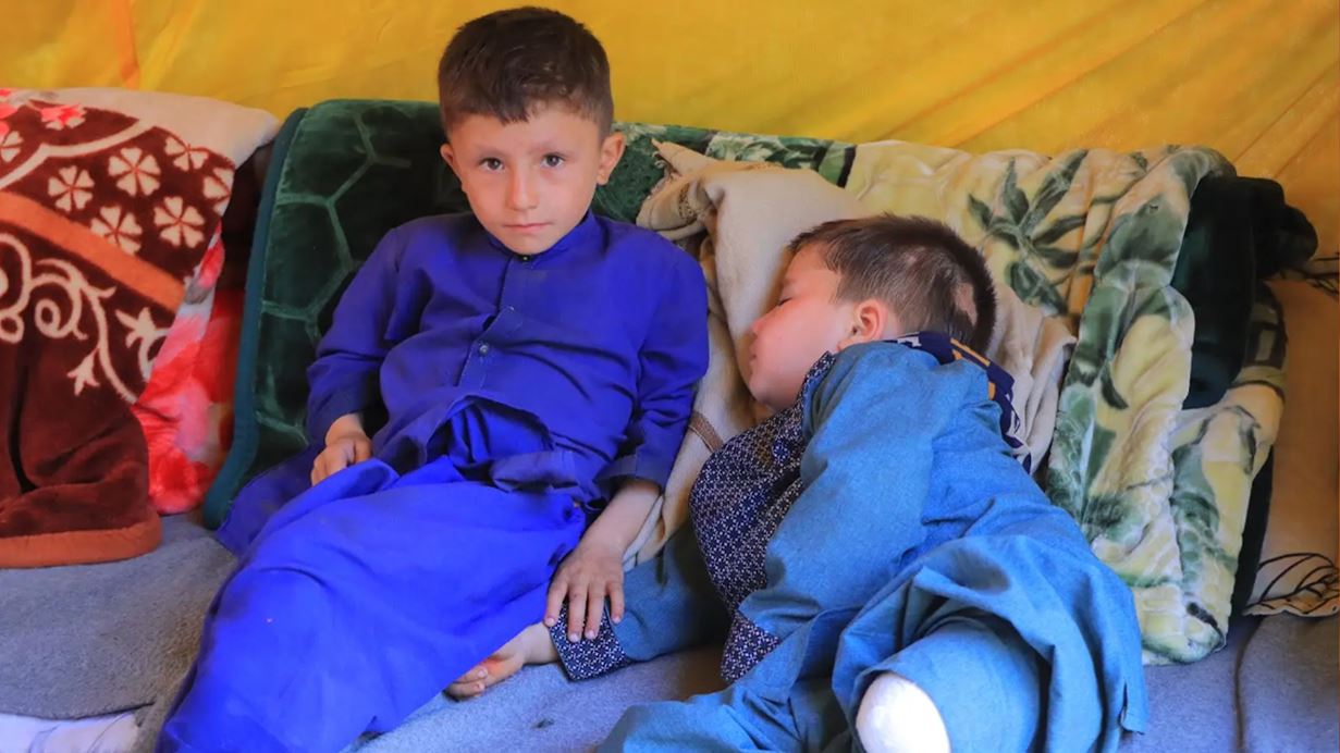 Two young boys rest on a blanket, one awake and alert, the other asleep.
