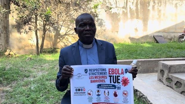 A person holding a sign about Ebola prevention, wearing a blazer and clerical collar.