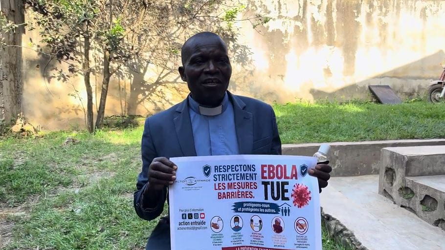 A person holding a sign about Ebola prevention, wearing a blazer and clerical collar.