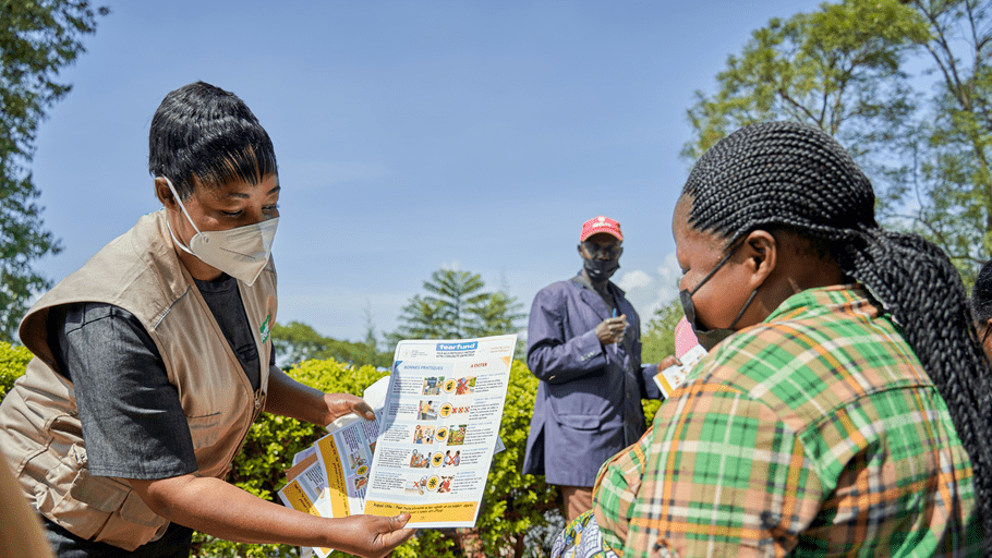 A community health worker wearing a face mask and beige vest hands an educational pamphlet to a woman outdoors.