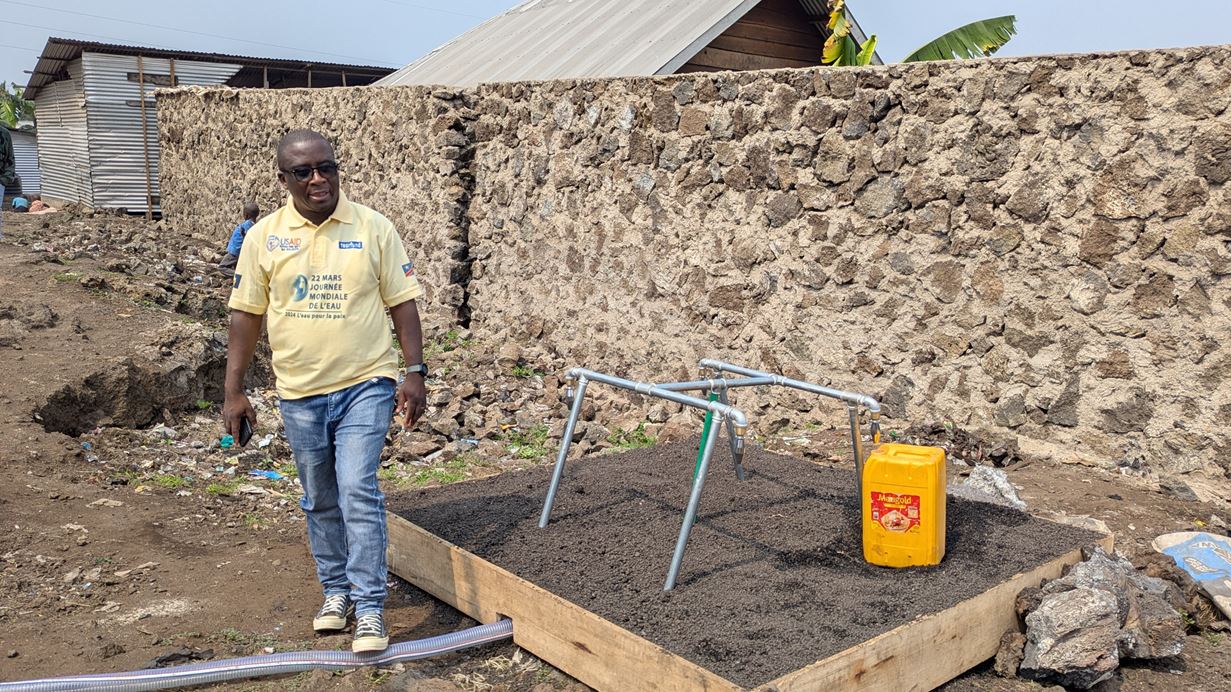 A man in a yellow Tearfund shirt and sunglasses stands next to a water tap installed by Tearfund in DRC