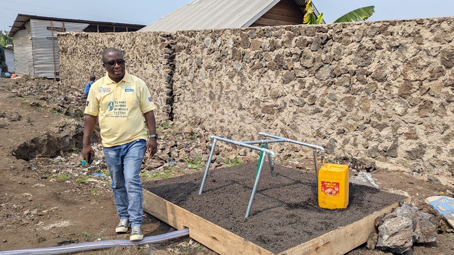 A man in a yellow Tearfund shirt and sunglasses stands next to a water tap installed by Tearfund in DRC
