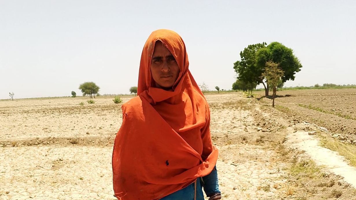 A woman in a bright headscarf stands on cracked, parched ground under a clear blue sky.
