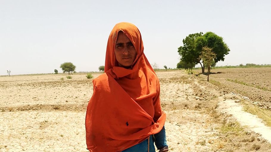 A woman in a bright headscarf stands on cracked, parched ground under a clear blue sky.