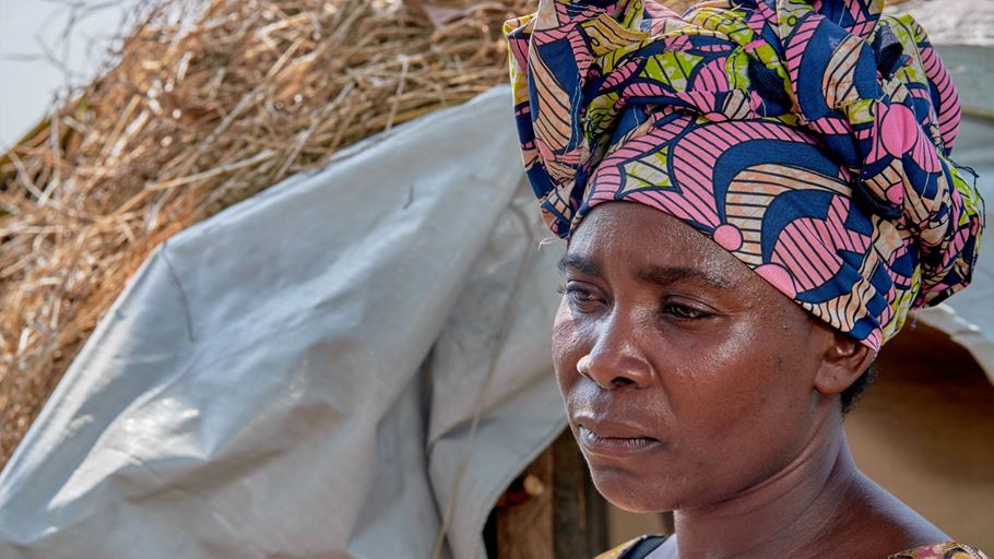 A woman looks to the side of the camera in front of a straw structure.